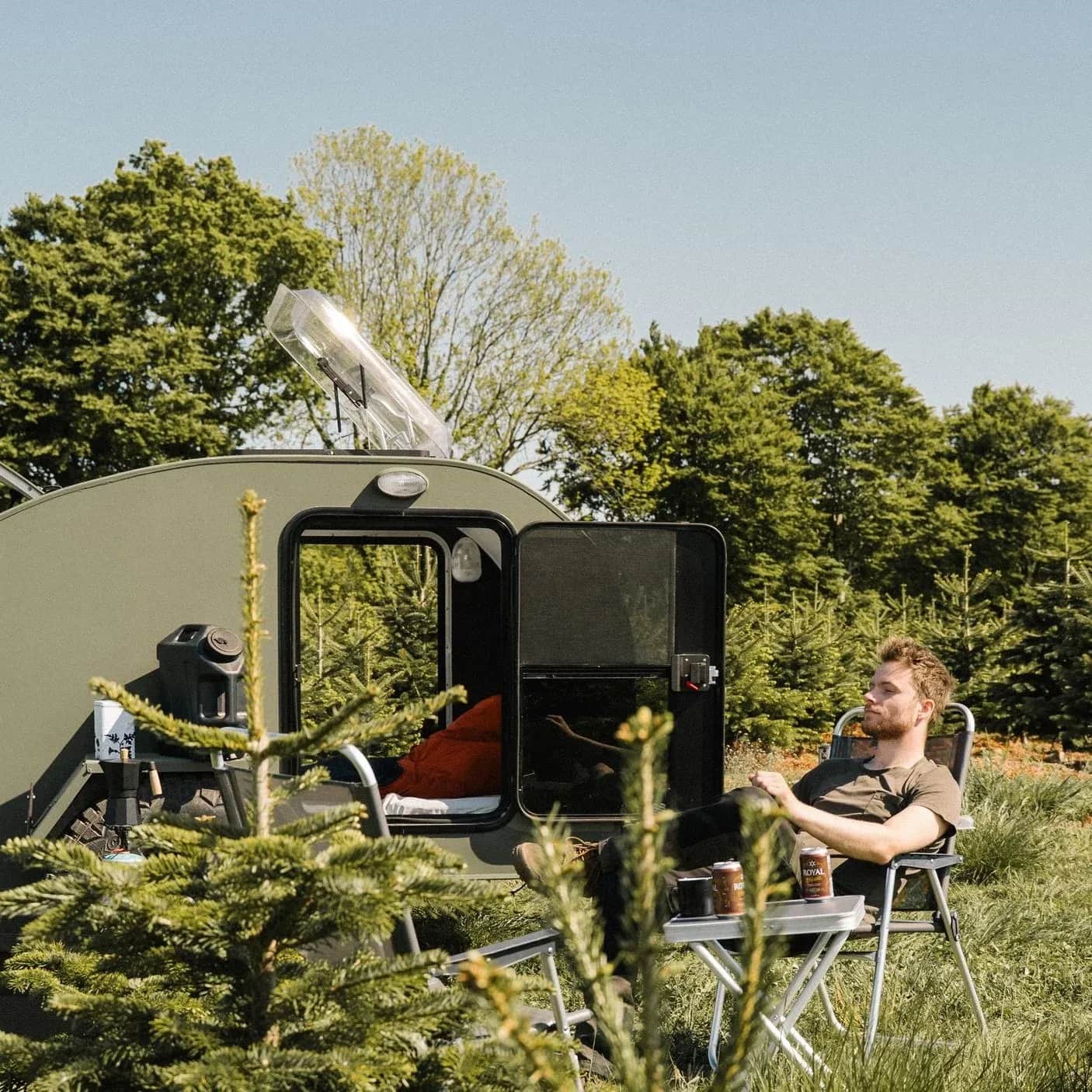 Person sidder i en stol ved en åben campingvogn mellem træer.