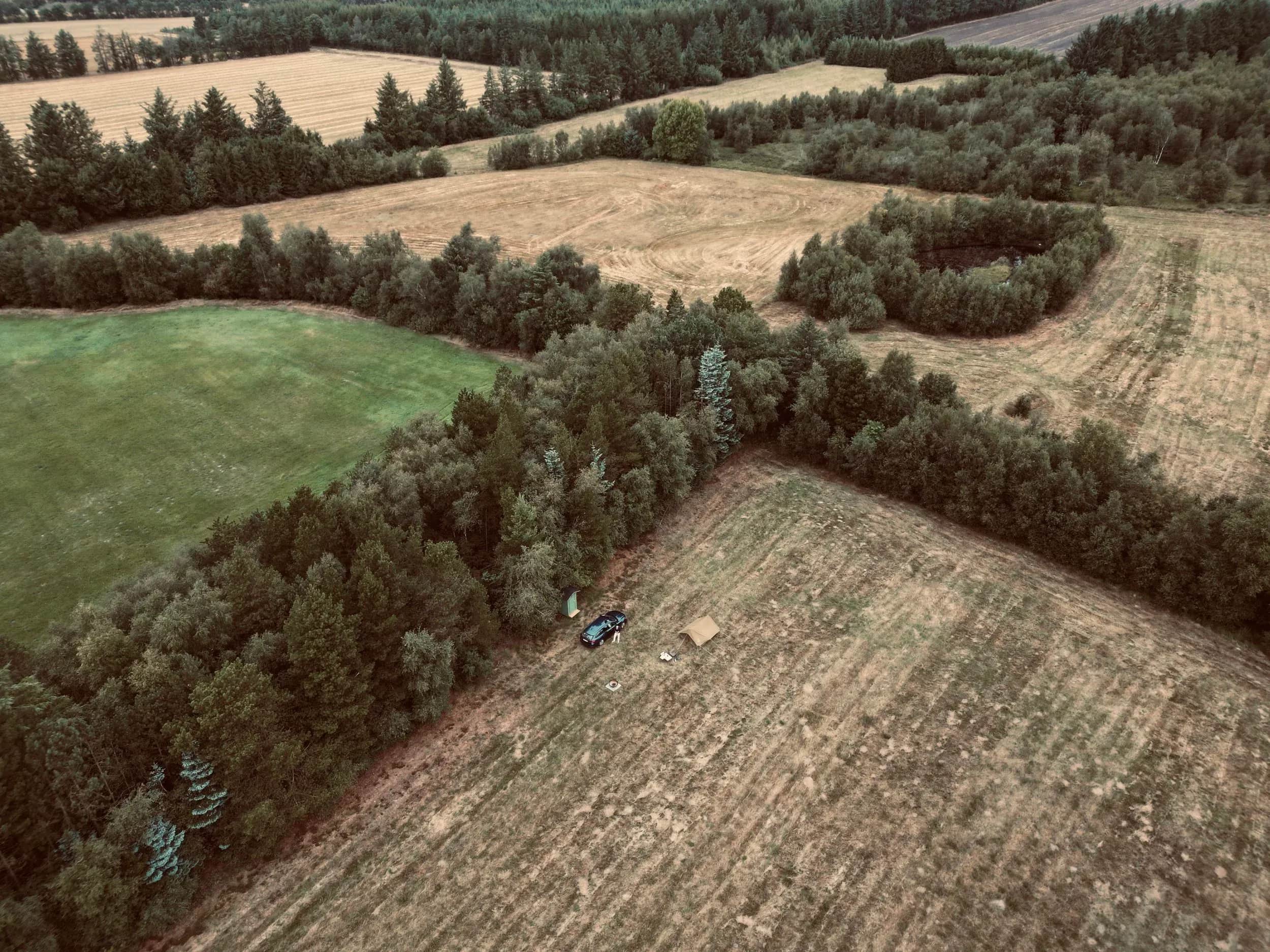 Luftfoto af en bil og et telt i et markhjørne ved skoven.