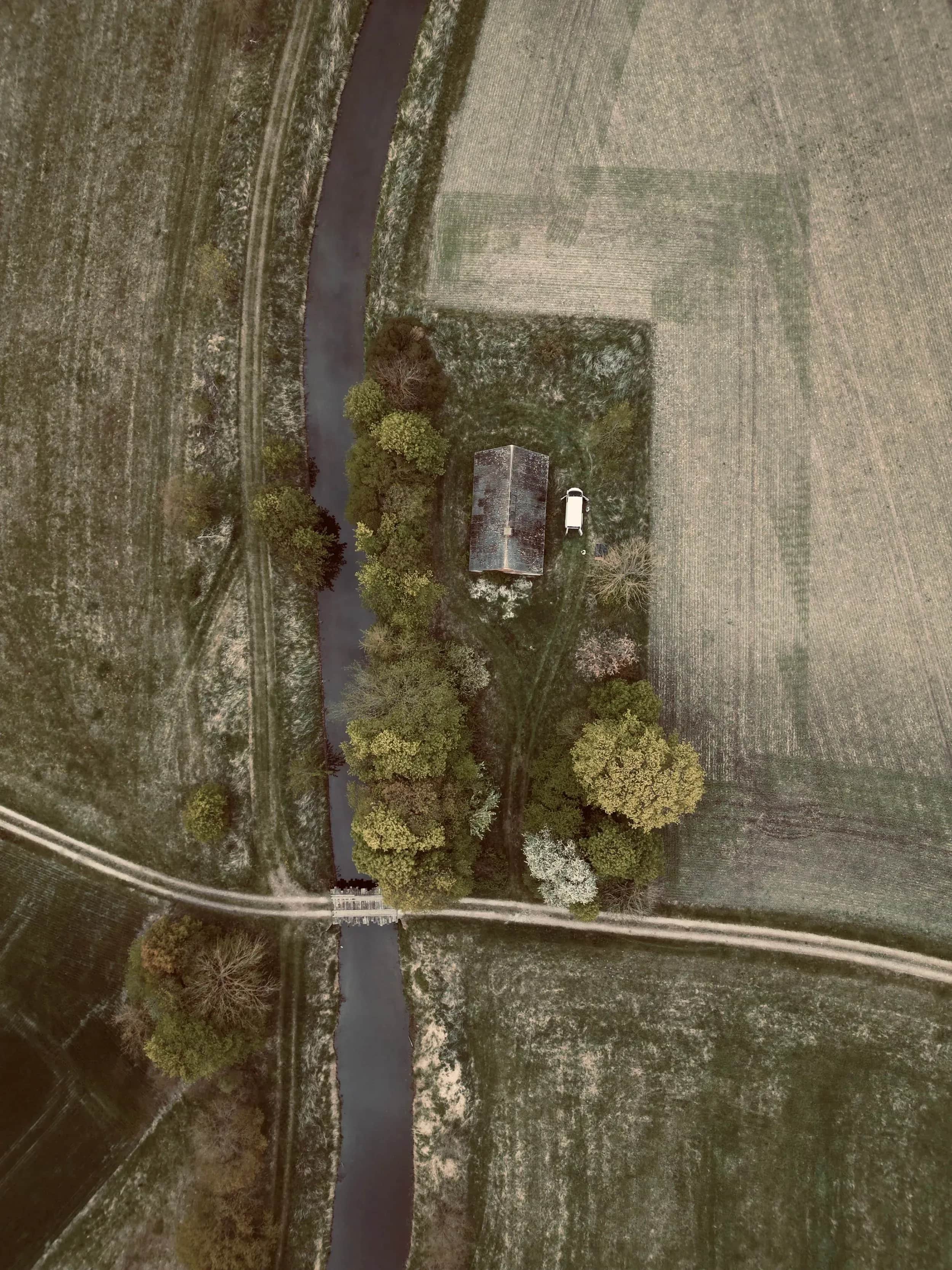 Luftfoto af en å, en vej og et lille hus mellem marker.