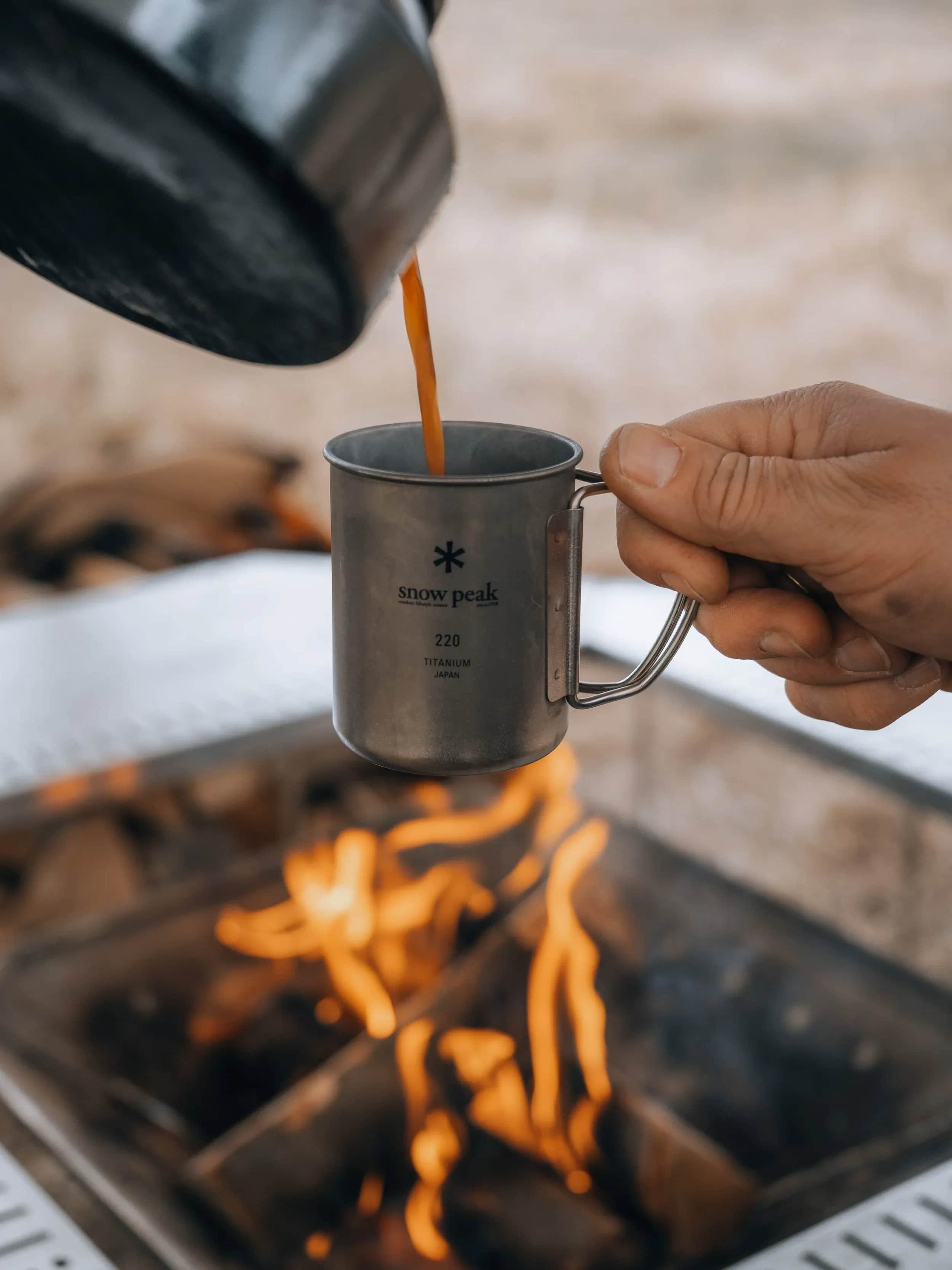 Kaffe hældes i et metalkrus over et bål.